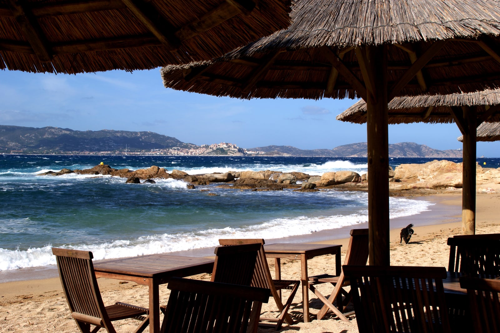 Strandbar mit Strohschirmen und Holztischen am Sandstrand, Blick übers Meer auf die Zitadelle von Calvi